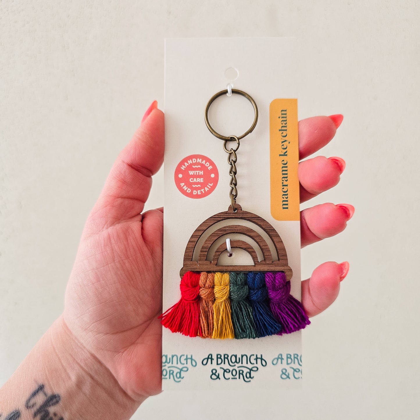 A hand holding a wooden keychain with a rainbow pattern and tassel, on a white background with a product tag.