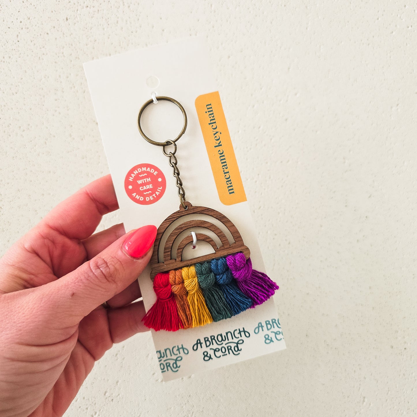 A hand holding a wooden keychain with a rainbow pattern and tassel, on a white background with a product tag.