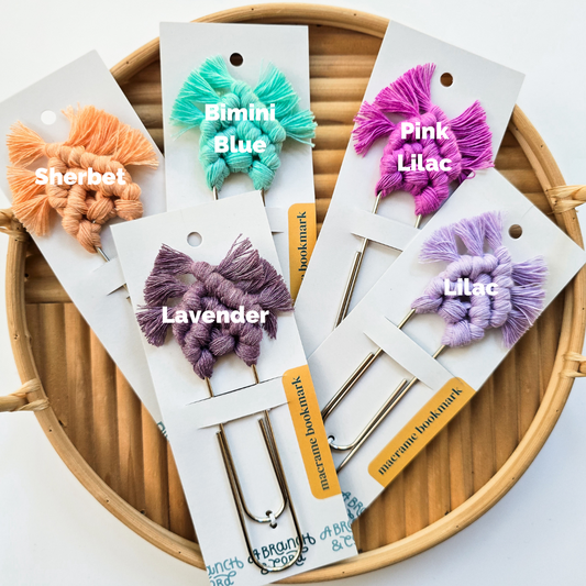A selection of handcrafted macrame bookmarks in various pastel colors, displayed on cards with a bamboo-like texture in the background.