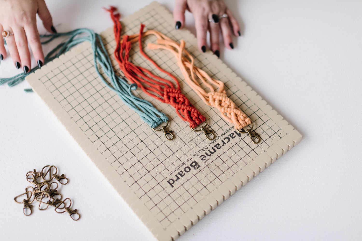 Colorful macrame keychains being made on a grid board with hands and a pile of bronze claps.