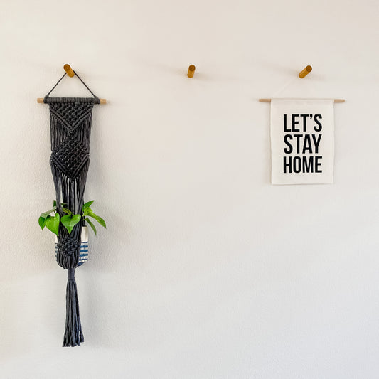 A macrame plant hanger in gray, hanging on a wall with a white sign that says 'LET'S STAY HOME' and a green plant inside.