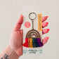A hand holding a wooden keychain with a rainbow pattern and tassel, on a white background with a product tag.