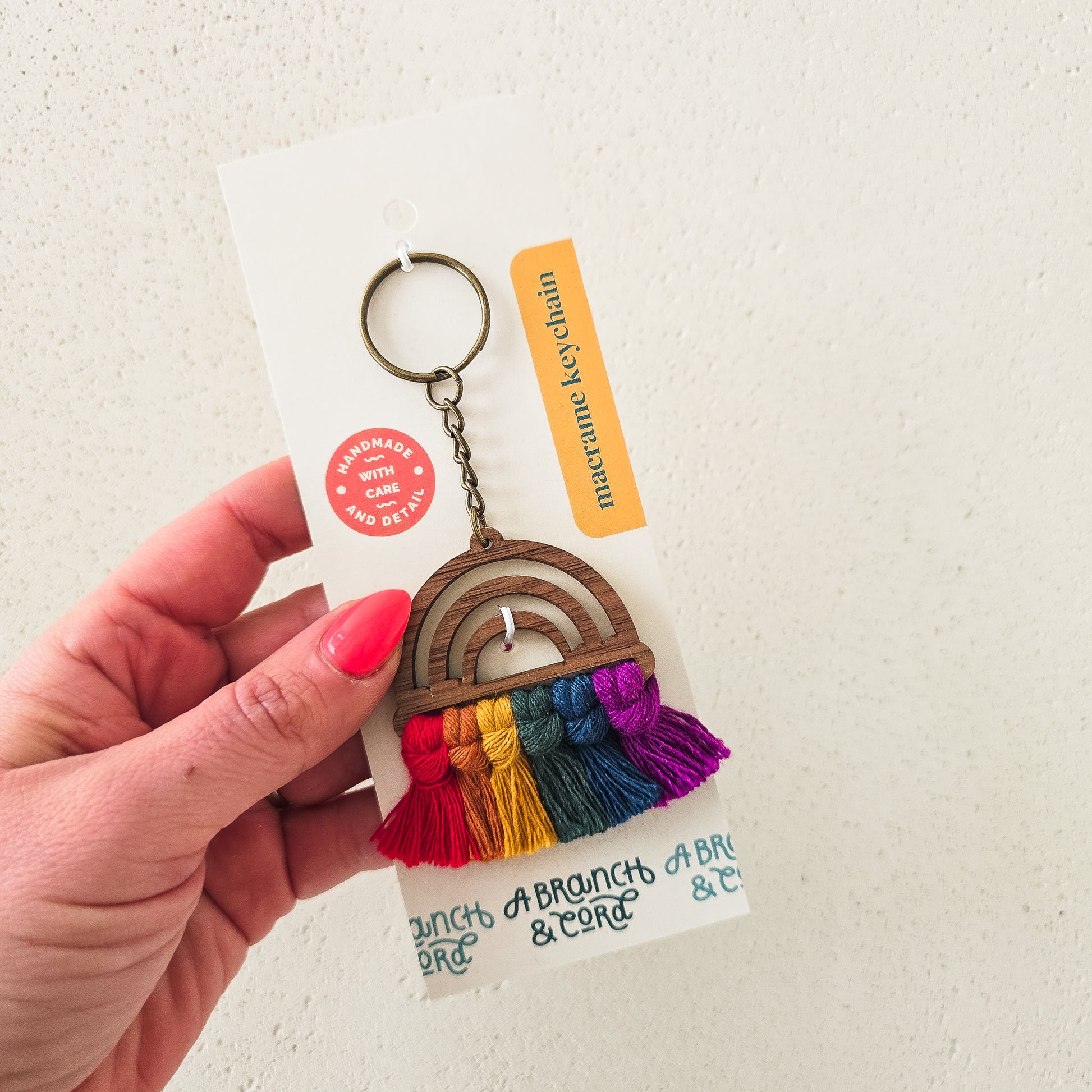 A hand holding a wooden keychain with a rainbow pattern and tassel, on a white background with a product tag.
