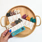 Hand holding a box of hair ties with a tray of colorful hair ties on a white background