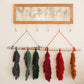 Colorful macrame feathers hanging from a wooden frame against a white wall.
