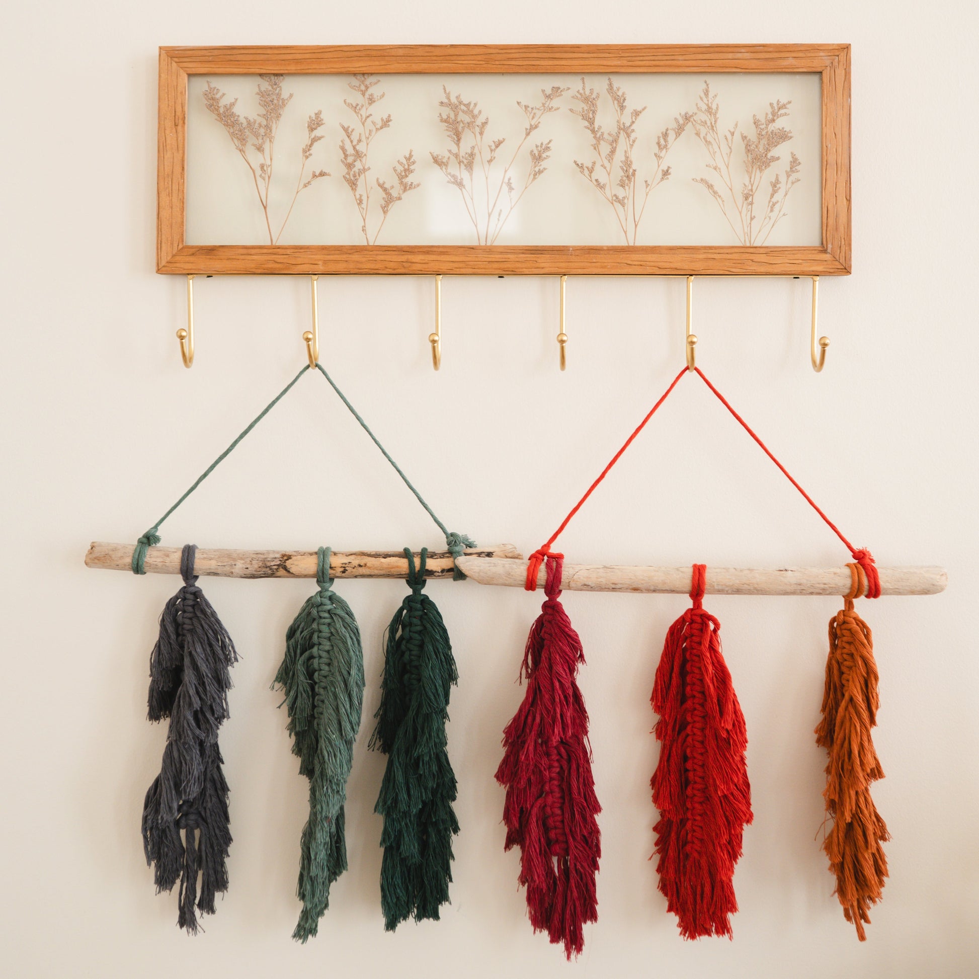 Colorful macrame feathers hanging from a wooden frame against a white wall.