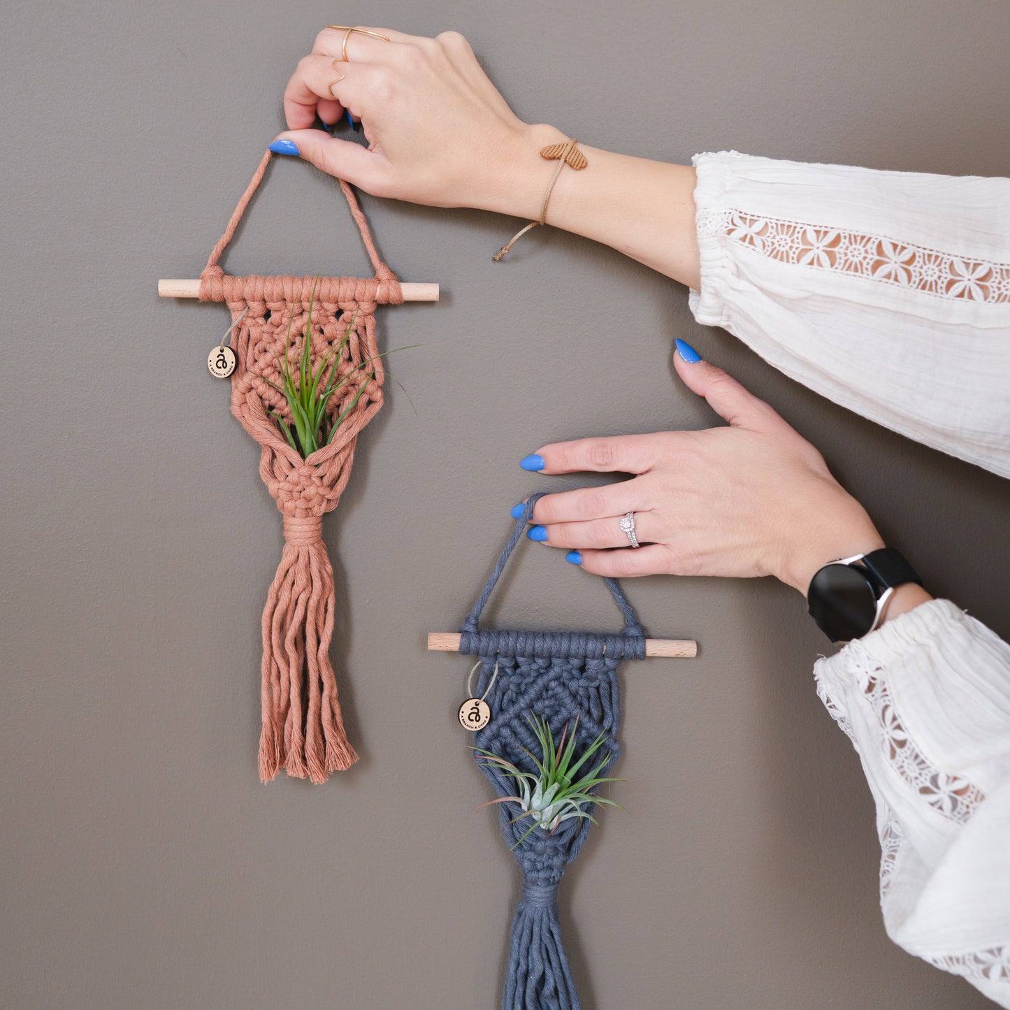 Two macrame wall hangings being held against a plain wall.