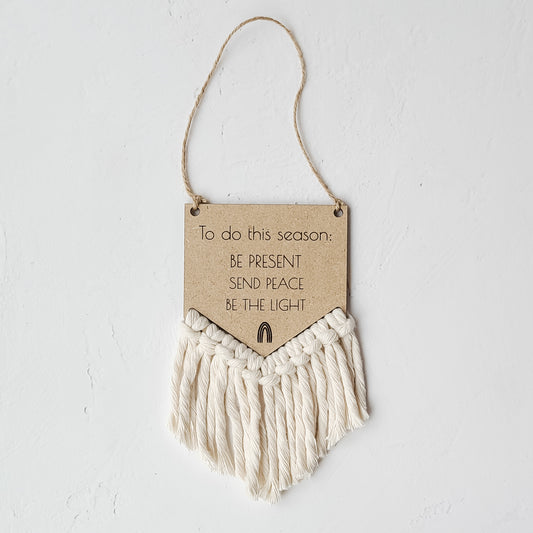 A round bamboo wall decor featuring a hanging macrame cord with a wooden sign that has text 'To do this season: BE PRESENT, SEND PEACE, BE THE LIGHT'.