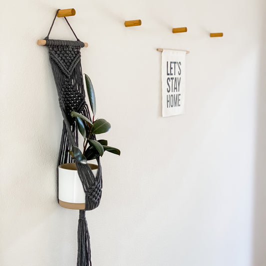 A macrame plant hanger in gray, hanging on a wall with a white sign that says 'LET'S STAY HOME' and a green plant inside.